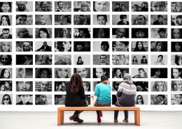 black and white photos of different faces covering a large wall, with three women on a bench looking at them