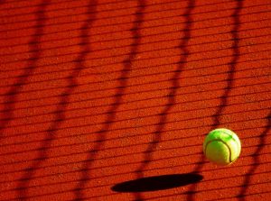 green tennis ball bouncing off of a red clay court with the shadow of the net across the court
