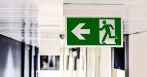 Green emergency exit graphic sign with human figure running through a door and an arrow pointing left