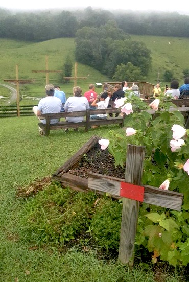 people sitting on benches on a hill facing away from camera and toward three wooden crosses with another wooden cross and flowers in the foreground