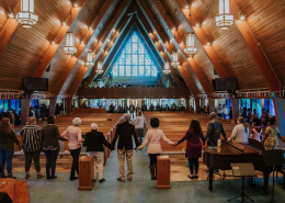 Group of people holding hands around an A-frame wooden church sanctuary