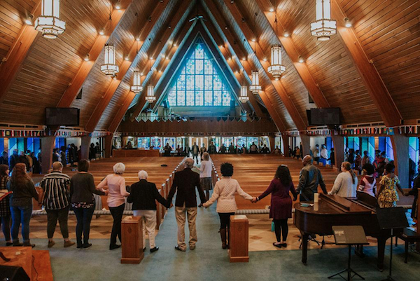 Group of people holding hands around an A-frame wooden church sanctuary