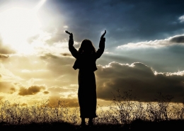 woman in silhouette with arms raised standing in grass in front of a sunset or sunrise